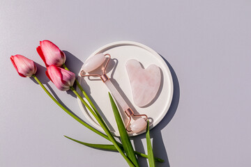 Rose quartz roller massager and scraper on a round ceramic tray with live tulips. top view. gray background. Natural care.