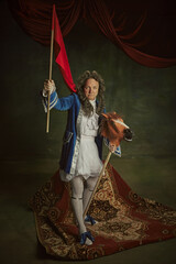 Elderly man wearing in baroque costume holding red flag, sitting on playful horse-toy against...