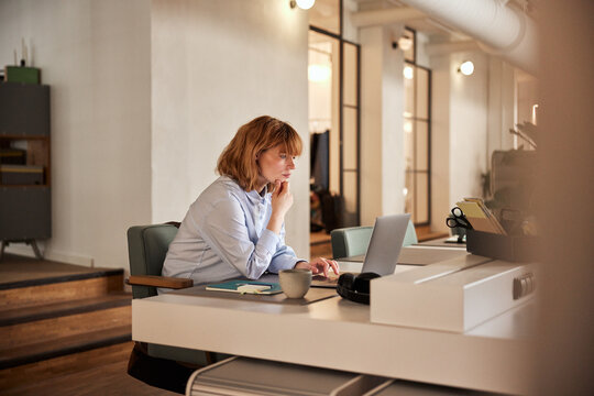 A business woman with red hair and casual work clothes, sitting focused and reading something on her laptop in the evening