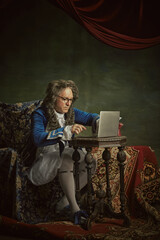 Man, dressed as king in classical attire and glasses sitting working online in modern laptop...
