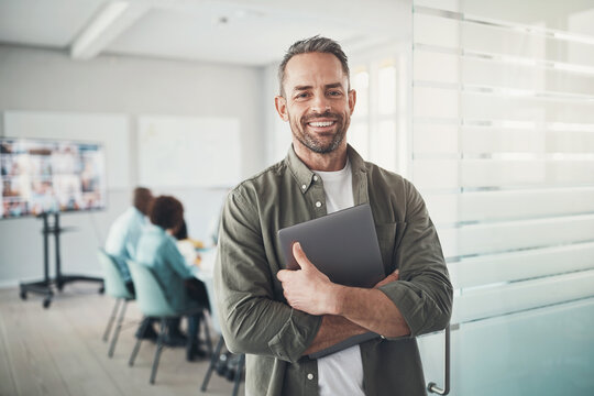 Smiling businessman standing with a laptop by a boardroom before meeting with diverse colleagues