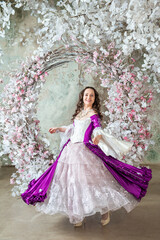 Happy beautiful woman in fantasy white and purple rococo style medieval dress dancing near decoration with white flowers