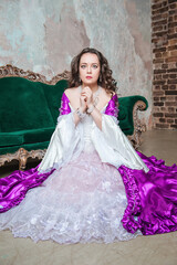Fototapeta premium Sad beautiful woman in fantasy white and purple rococo style medieval dress sitting on the floor near sofa and praying