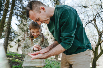 Happy girl examining bug on father's hand with magnifying glass in forest