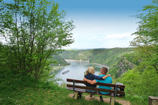 Hiking couple with arms around sitting on bench by view of Loreley Rock in St. Goarshausen, Rhineland Palatinate, Germany