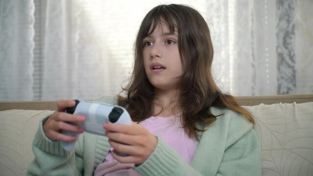 11 years old child holding gaming console and playing on a sofa. Portrait of pre-teenage girl with video game remote control playing a video game, holding joystick, smiling. Children's Leisure And