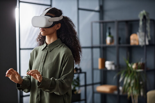 Medium shot of modern young biracial woman wearing VR headset standing in living room typing on virtual keyboard in air, copy space