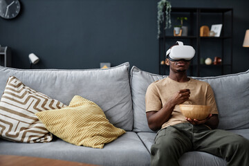 Young African American man wearing VR headset sitting on sofa in living room at home eating snacks...