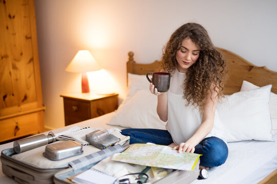 Beautiful woman planning summer vacation abroad, sitting on bed, packing suitcase. Solo traveler looking at map, drinking coffee.