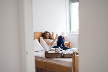 Beautiful woman lying on bed next to packed suitcase. Solo traveller preparing for trip.