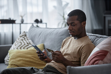 Young African American man sitting on sofa in living room paying for goods online using app on...