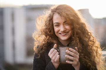 Beautiful woman with curly hair standing on terrace, drinking coffee during cold spring day. Morning sun, hair blowing in wind.