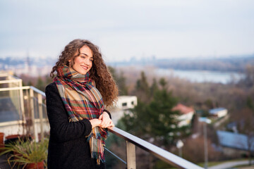 Beautiful woman with curly hair standing on terrace, enjoying cold autumn morning, sunny day....