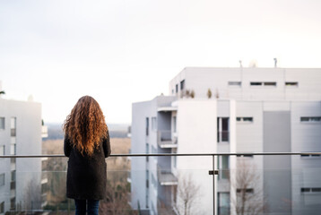 Naklejka premium Rear view of woman with curly hair standing on terrace, enjoying cold autumn morning, sunny day. Speaking positive affirmations.