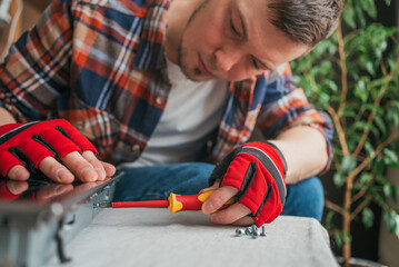 Focused man wearing plaid shirt and red gloves uses yellow screwdriver to assemble furniture indoors, with screws and parts laid out beside him.