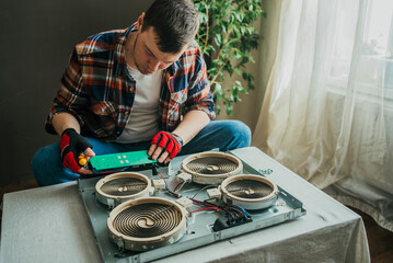 Concentrated technician in plaid shirt and red gloves repairs electric stove, carefully examining circuit board over disassembled appliance.