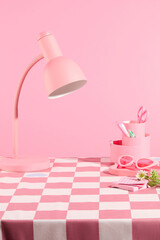 A table with checkered pink table cloth over pastel pink background, containing a lamp and some pink accessories above. Blank space for displaying product, front view