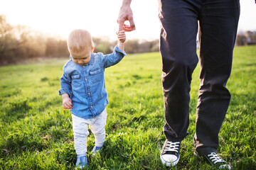 Portrait of father holding little toddler boy by hand, walking in spring nature. First steps for...