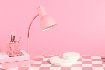 Frontal shot photo with a table has checkered pattern containing blank podium in center for displaying product next to a lamp with pink color and some makeup utensils placed on top of a few books