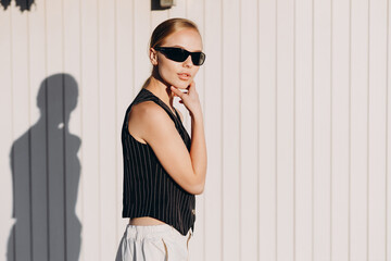 A young stylish girl with bun hair and black sunglasses on her eyes, wearing a black vest and white...