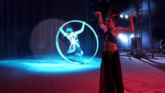 Gymnastics on wheels performed by a young man on the circus stage, a girl dancing in the foreground, a circus performance with smoke and colorful spotlights.A circus performer rotates on a cyr wheel.