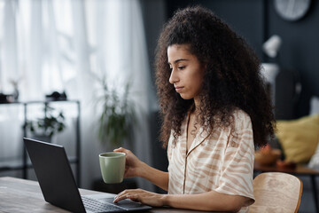 Medium shot of young biracial woman sitting at desk in living room in morning surfing Internet on...