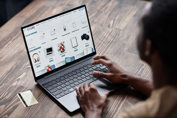 High angle view of unrecognizable Black man sitting at wooden desk choosing goods in online shop on laptop