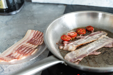 Close-up of sauteed bacon and tomatoes in a pan next to a plate of bacon. Delicious breakfast. Selected focus