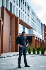 A man in a black shirt and jeans stands in front of a building. The building is a large, modern structure with a white facade.