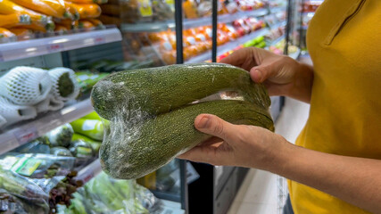 Person in a yellow shirt selecting a large zucchini in a grocery store, conceptually related to...