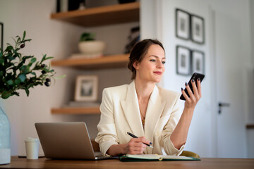 Attractive woman sitting at desk and using smartphone and laptop at home