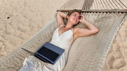 Smiling caucasian woman relaxing on a hammock with a laptop, embodying remote work and leisure...