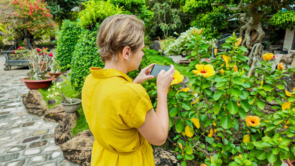 A young Caucasian child in a yellow shirt takes a photograph of vibrant flowers with a smartphone in a lush garden, symbolizing spring and Earth Day