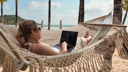 Caucasian woman using a laptop on a tropical beach with palm trees, embodying leisure technology use and summer vacations, freelancer, digital nomad