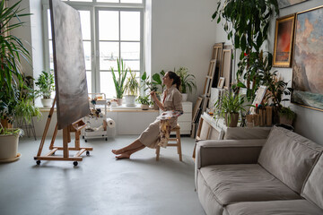 Perfectionist woman artist critically evaluates own painting, sitting on chair, looking for flaws,...