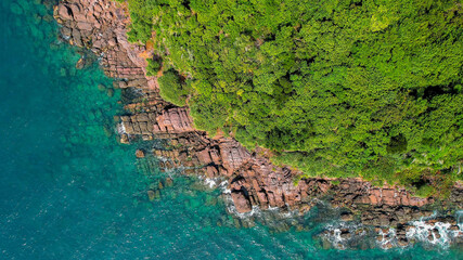 Aerial view of a rugged coastline with red rock formations and lush greenery meeting the turquoise...