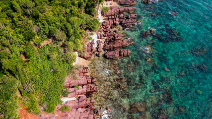 Aerial view of a rugged coastline with red rock formations and lush greenery meeting the turquoise...