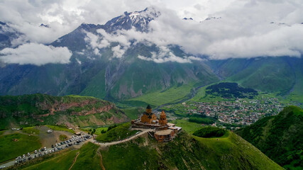Obraz premium Aerial view of a historic monastery perched on a hilltop overlooking a valley village with snow-capped mountains and cloudy skies, possibly Georgia