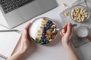 Woman eating tasty granola at white wooden table with laptop, top view
