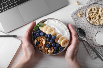 Woman holding bowl of tasty granola at white wooden table with laptop, top view