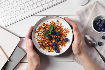 Woman holding bowl of tasty granola with blueberries at white wooden table with computer keyboard,...