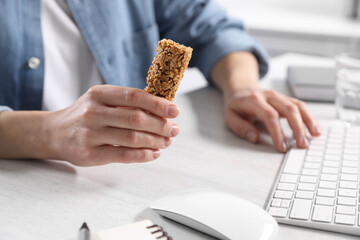 Woman holding tasty granola bar working with computer at light table in office, closeup