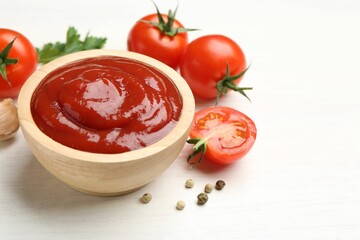 Delicious ketchup in bowl, peppercorns and tomatoes on white wooden table, closeup. Space for text