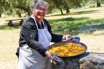 woman chef with white apron showing her typical Spanish paella cooked on the fire in the countryside