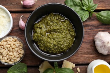 Tasty pesto sauce in bowl, basil, pine nuts, cheese, garlic and oil on wooden table, flat lay