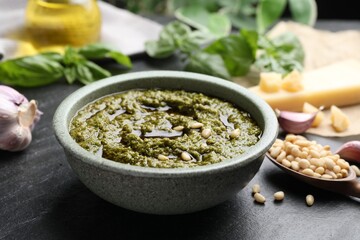 Tasty pesto sauce in bowl, pine nuts and garlic on black table, closeup