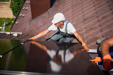 Worker building solar panel system on rooftop of house. Man engineer in helmet installing photovoltaic solar module outdoors. Alternative, green and renewable energy generation concept.