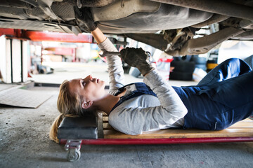 Female auto mechanic lying on mechanic creeper under car, inspecting and repairing vehicle....