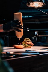 Vertical closeup of a hand with a black glove adding mayonnaise to the burger.