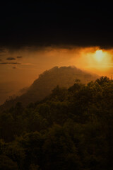 Vertical shot of a forested mountain in misty and stormy weather with a sunset sky in the background